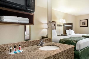 Quarto de hotel com lavatório de bancada de granito, um espelho, televisão sobre o balcão, cama com roupa de cama verde, mesa de cabeceira com luminária e um quadro na parede.