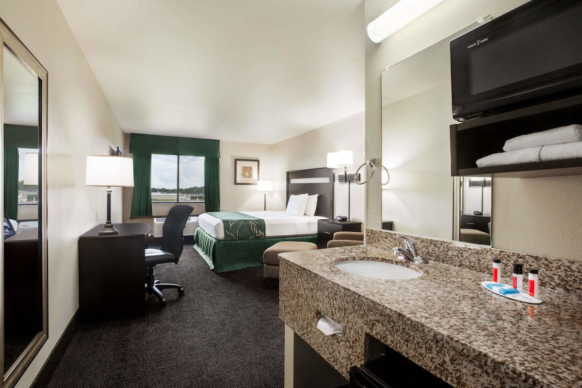 A hotel room with a bed, desk, chair, and a bathroom with a sink and mirror.