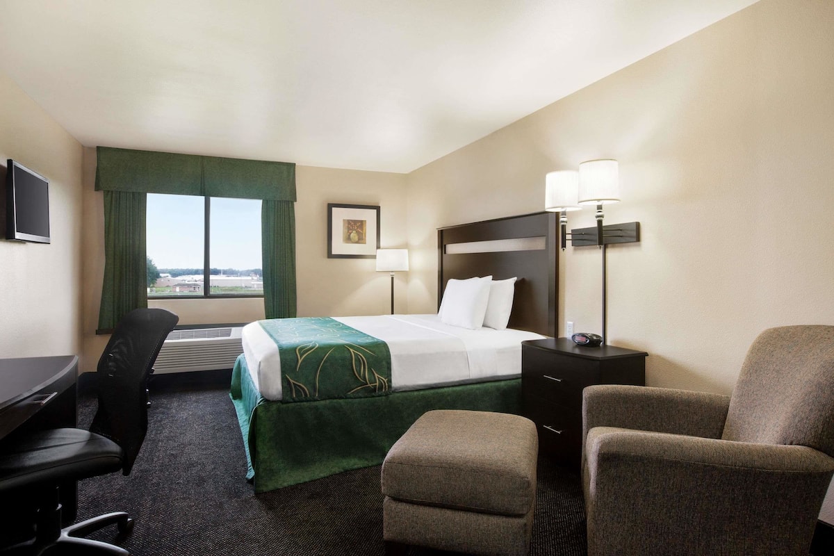 A hotel room with a bed, a desk, two chairs, a nightstand, a lamp, and a window with a view.
