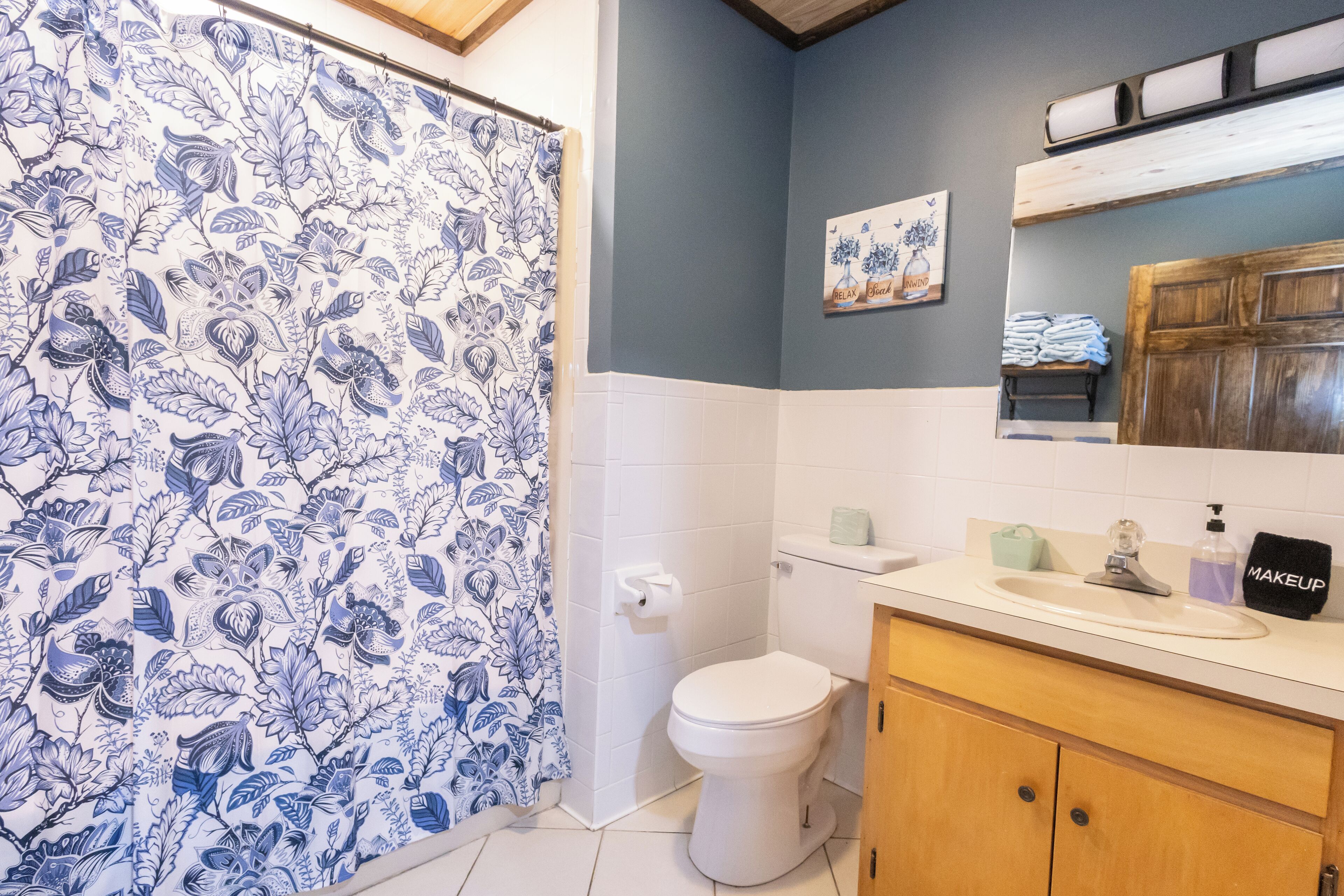 Un bany amb cortina de bany floral, un vàter, un lavabo amb mirall i un penjallot de tovalloles.