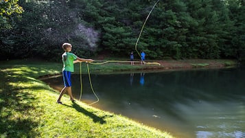 A person fishing by a calm pond surrounded by dense trees.