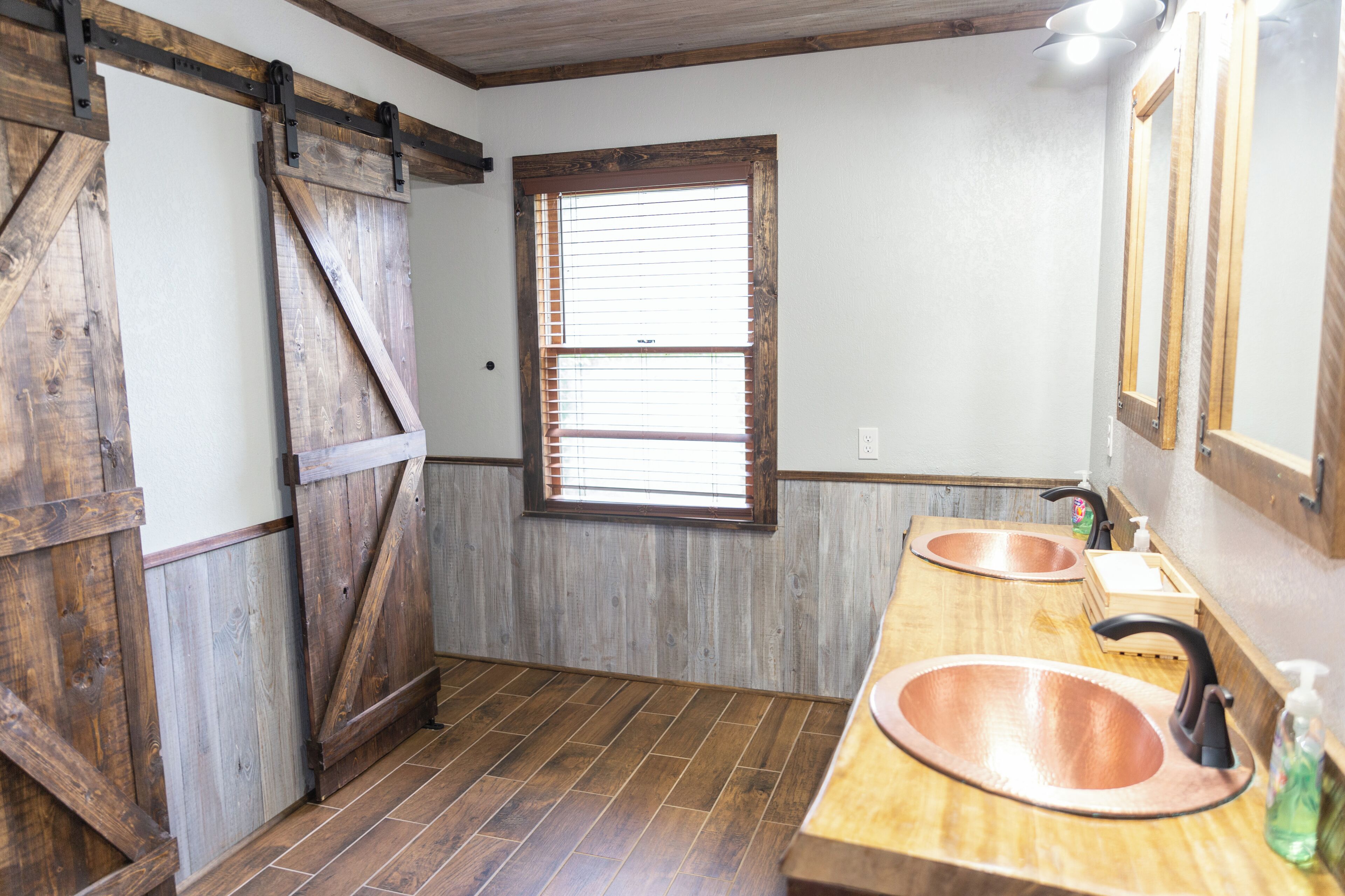 A rustic bathroom with wooden walls, a large window with blinds, and two copper sinks.
