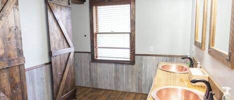 A rustic bathroom with wooden walls, a large window with blinds, and two copper sinks.