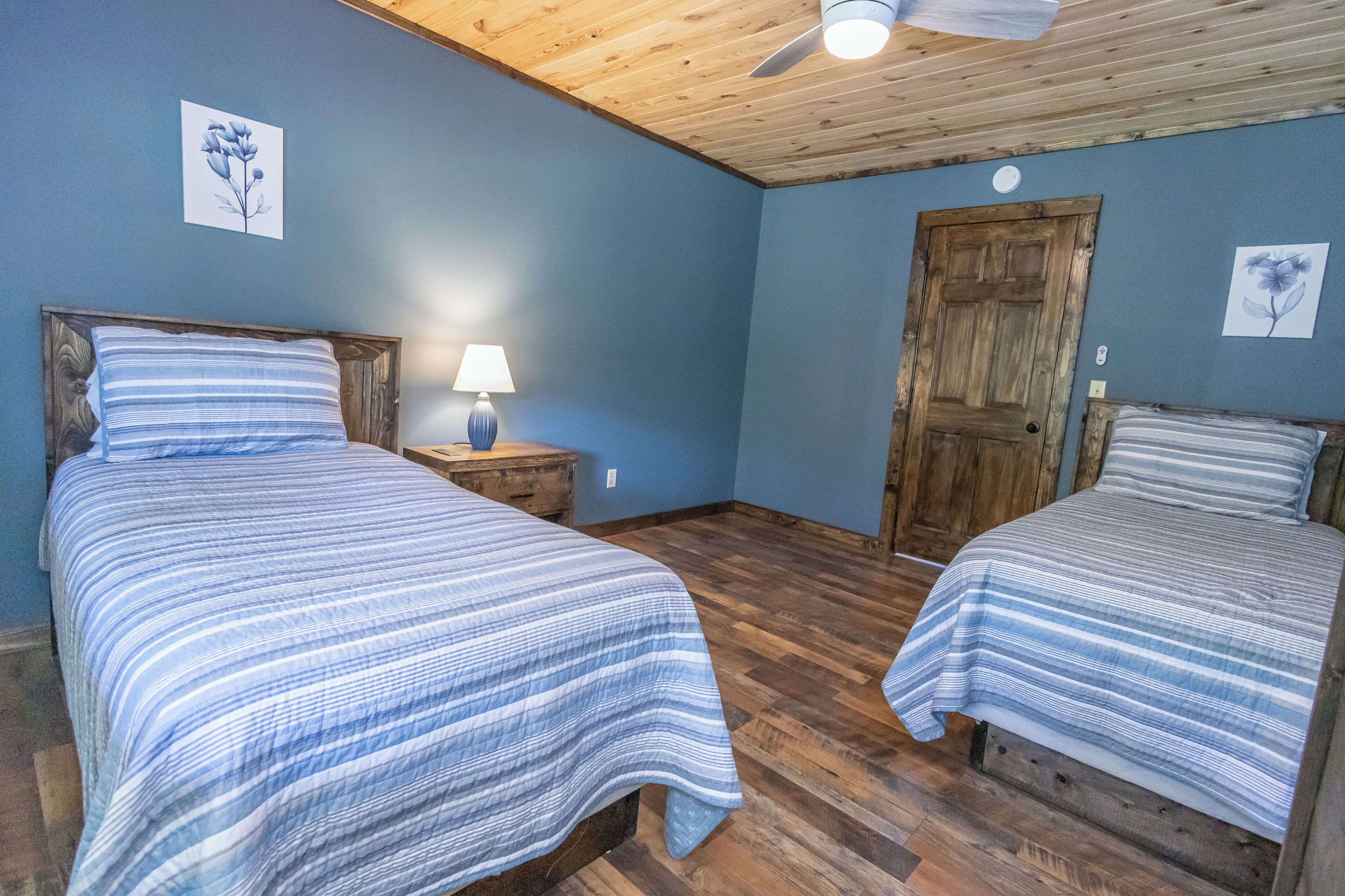 Ein Zimmer mit zwei Betten, Holzboden, Deckenventilator und einer Tür.