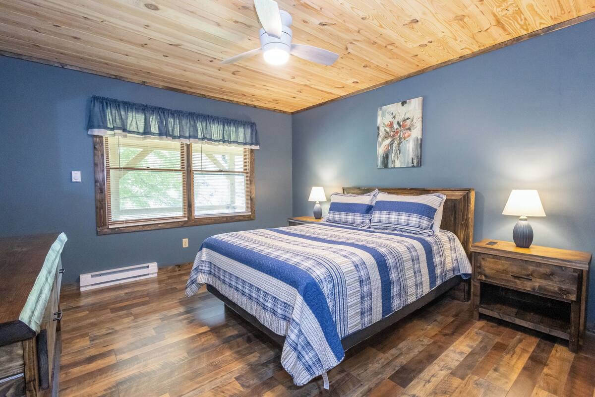 Une chambre avec un plafond en bois, un lit recouvert d’une courtepointe à rayures bleues et blanches, deux lampes de chevet et une fenêtre agrémentée d’une lambrequin bleue.