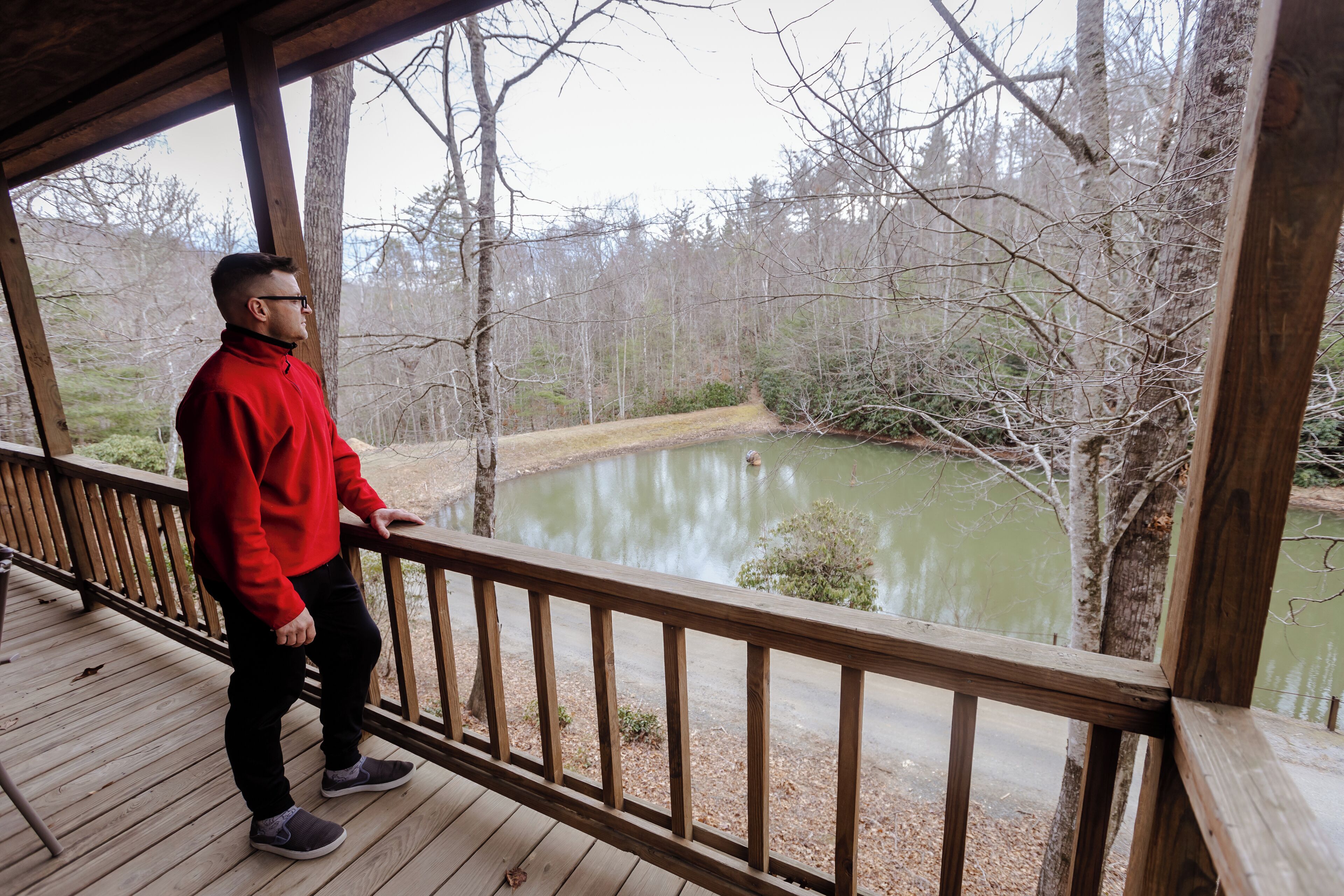A man standing on a wooden deck overlooking a pond surrounded by trees.