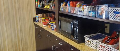 A kitchen with a microwave, shelves stocked with snacks, and storage cabinets.