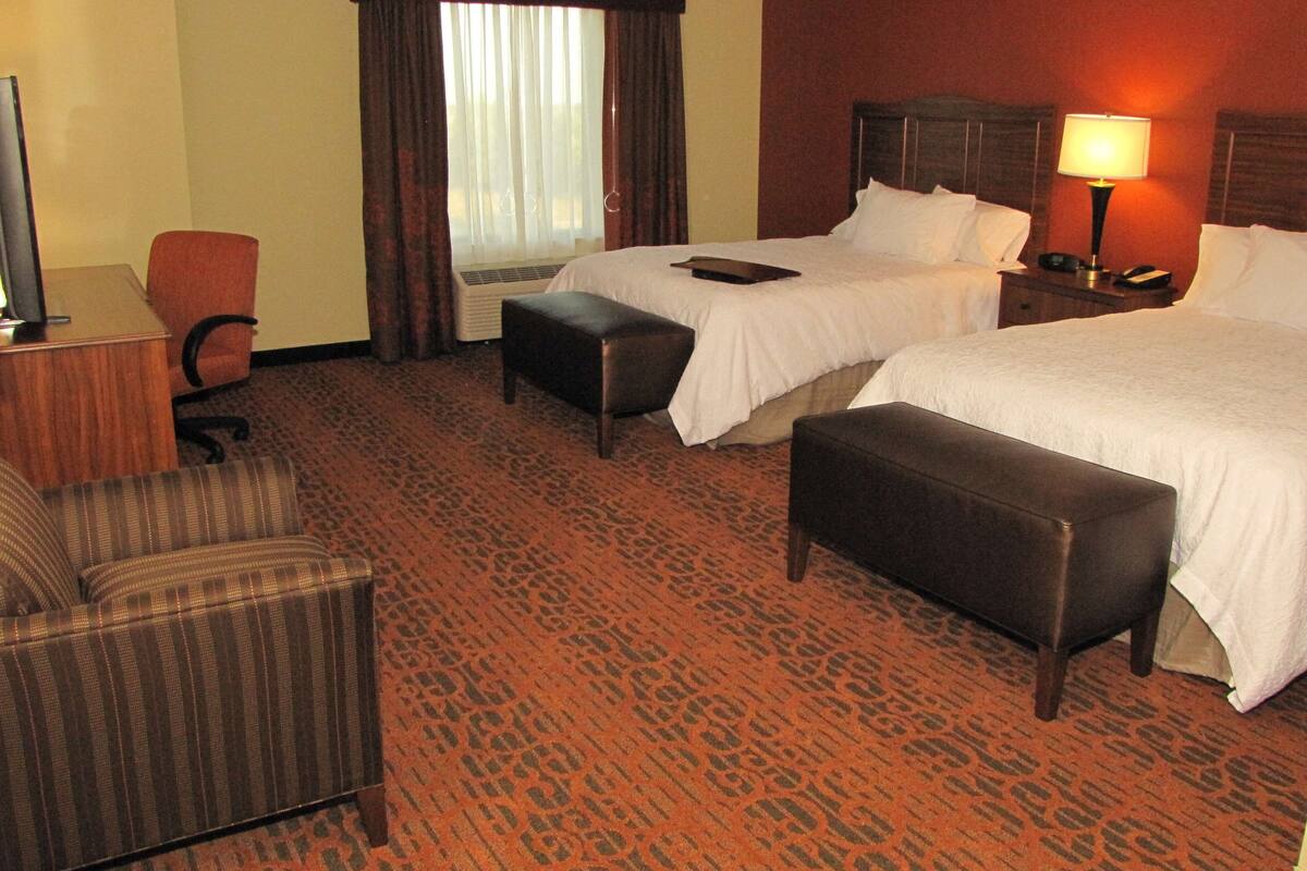 A hotel room with two beds, a desk, a chair, and a large window with curtains.