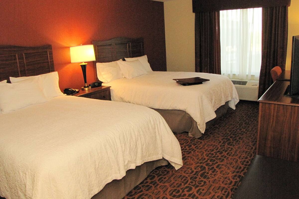 A hotel room with two beds, a nightstand with a lamp, a desk with a laptop, and a large window with curtains.