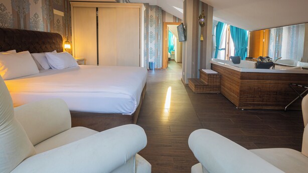 Une chambre d’hôtel avec un grand lit, un coin salon avec deux fauteuils, un bureau en bois et une salle de bain visible à travers une porte ouverte.