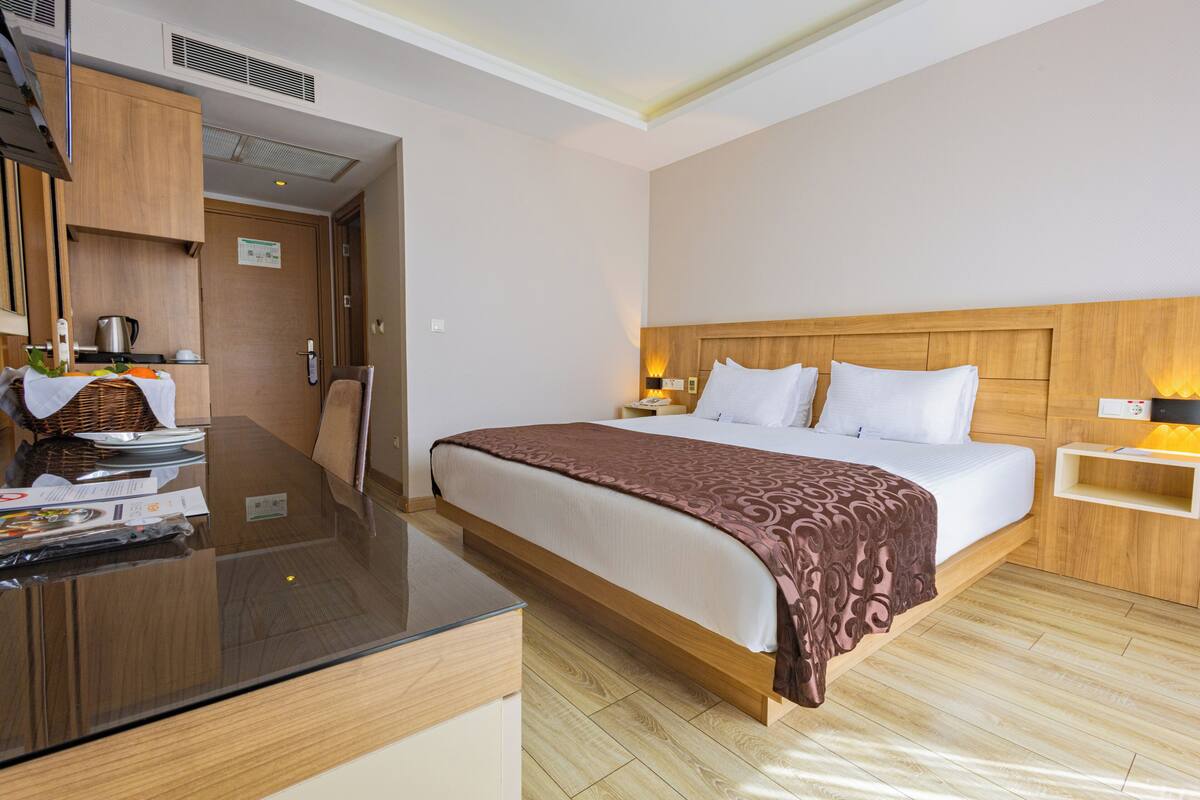 Habitación de hotel con cama, mesita de noche, escritorio con silla, armario de madera y zona de kitchenette.