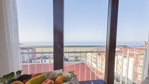 Une table avec un panier de fruits et un ensemble de couverts, donnant sur une vue côtière avec des bâtiments et une rue aux toits rouges.