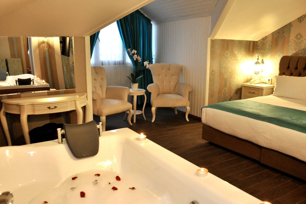 Habitación de hotel con una bañera grande, una cama, un escritorio con espejo, dos sillones y una mesita de noche con lámpara.