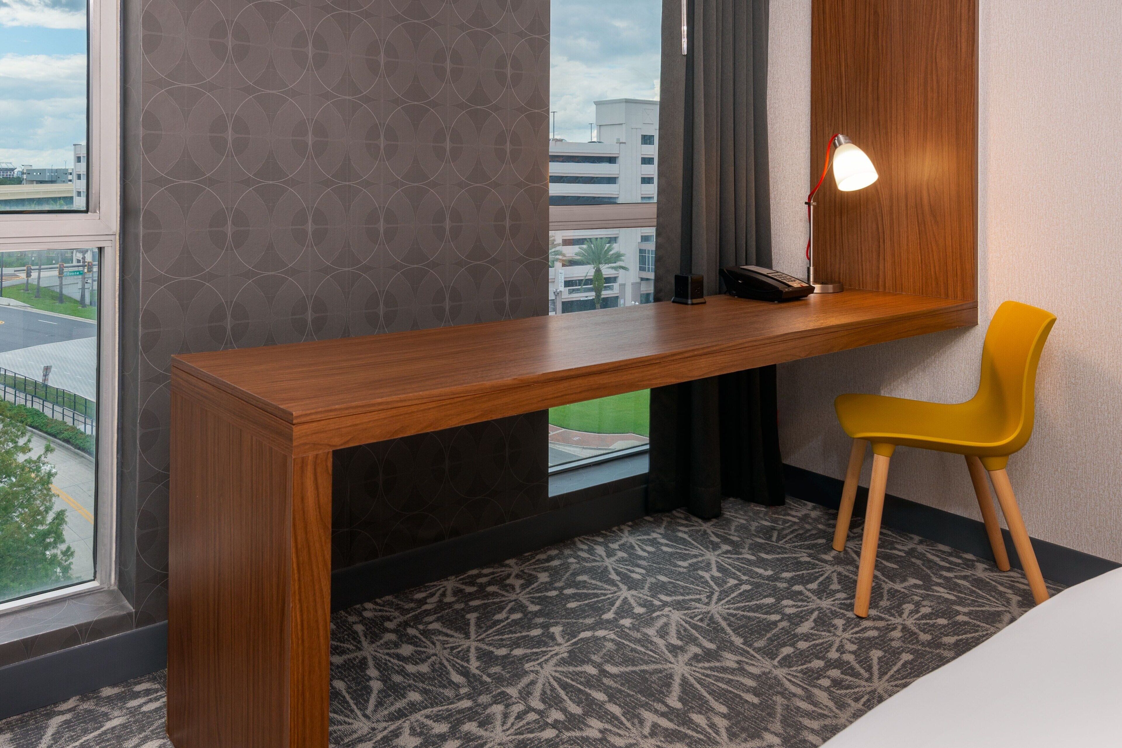 Habitación de hotel con un escritorio de madera, una silla amarilla, una lámpara y vistas de la ciudad a través de la ventana.