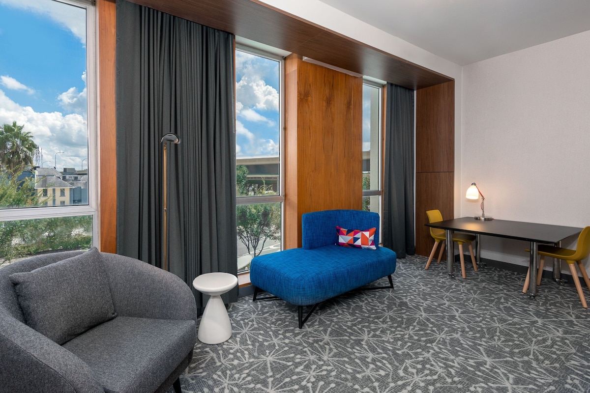 A hotel room with a grey armchair, a blue sofa, a small white side table, a dining table with yellow chairs, and a lamp.