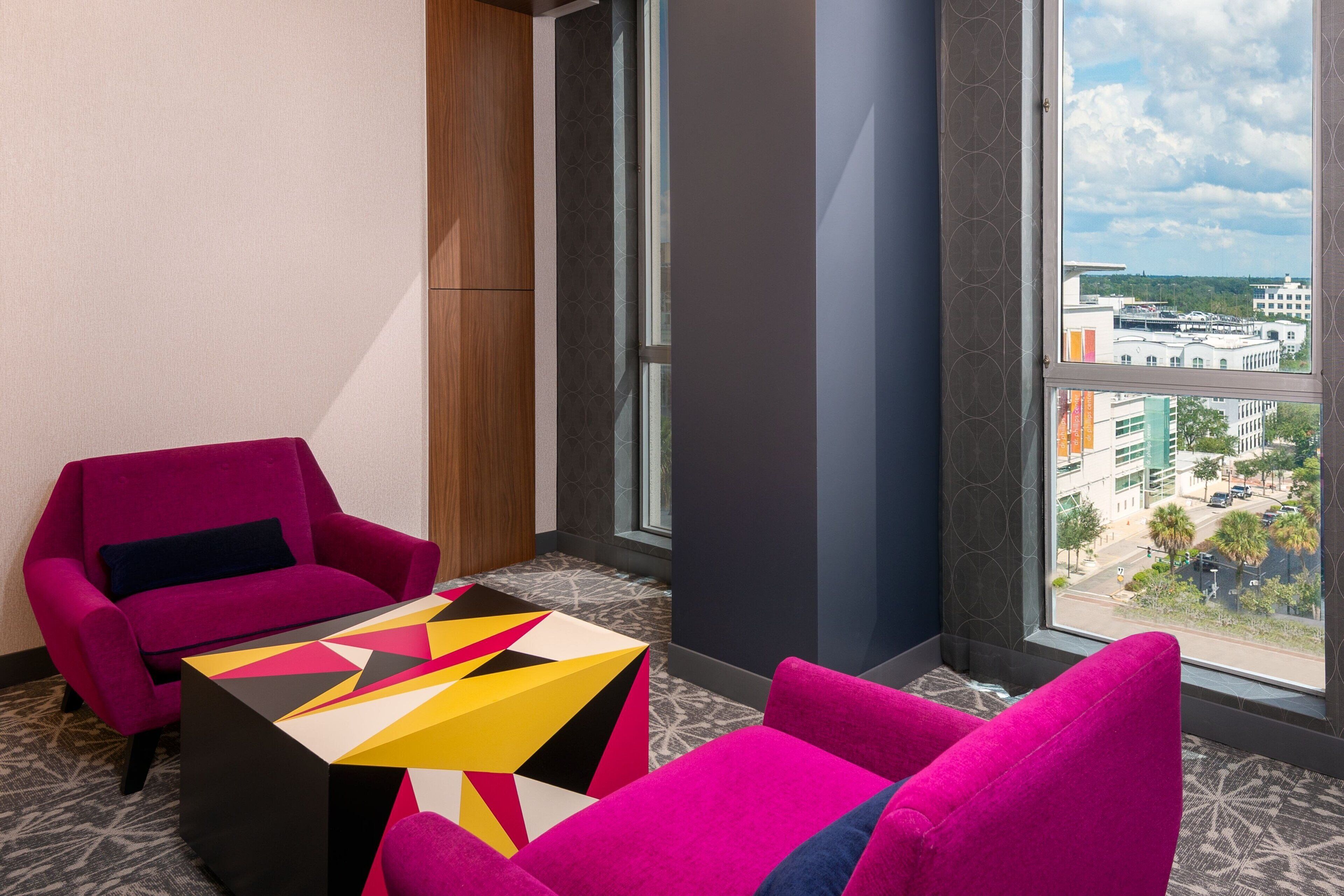 Ein modernes Hotelzimmer mit einem großen Fenster, einem leuchtend pinken Sessel, einem farbenfrohen geometrischen Couchtisch und einem gemusterten Teppich.
