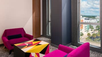 Ein modernes Hotelzimmer mit einem großen Fenster, einem leuchtend pinken Sessel, einem farbenfrohen geometrischen Couchtisch und einem gemusterten Teppich.