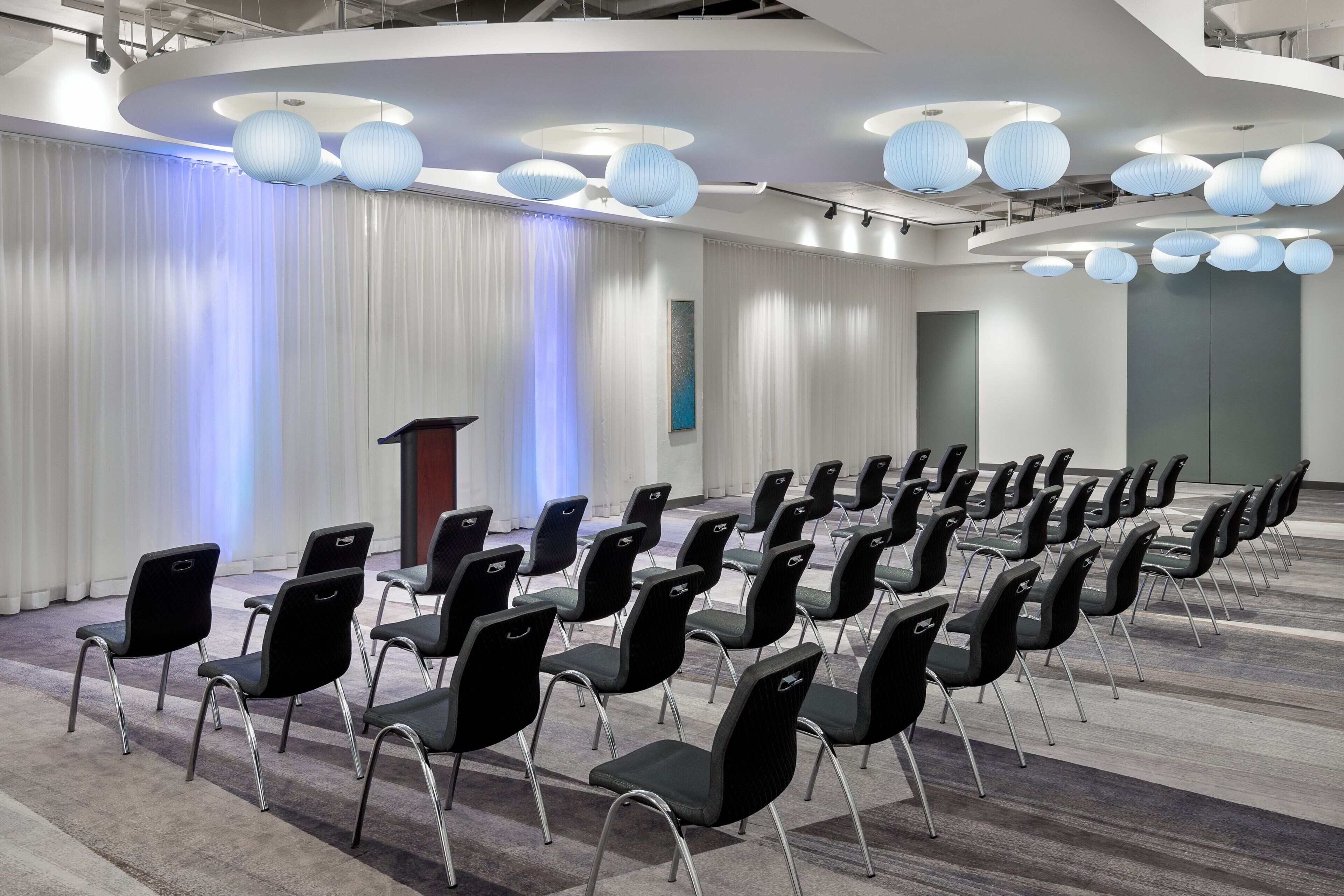 A conference room with rows of black chairs, a podium, and blue paper lanterns on the ceiling.