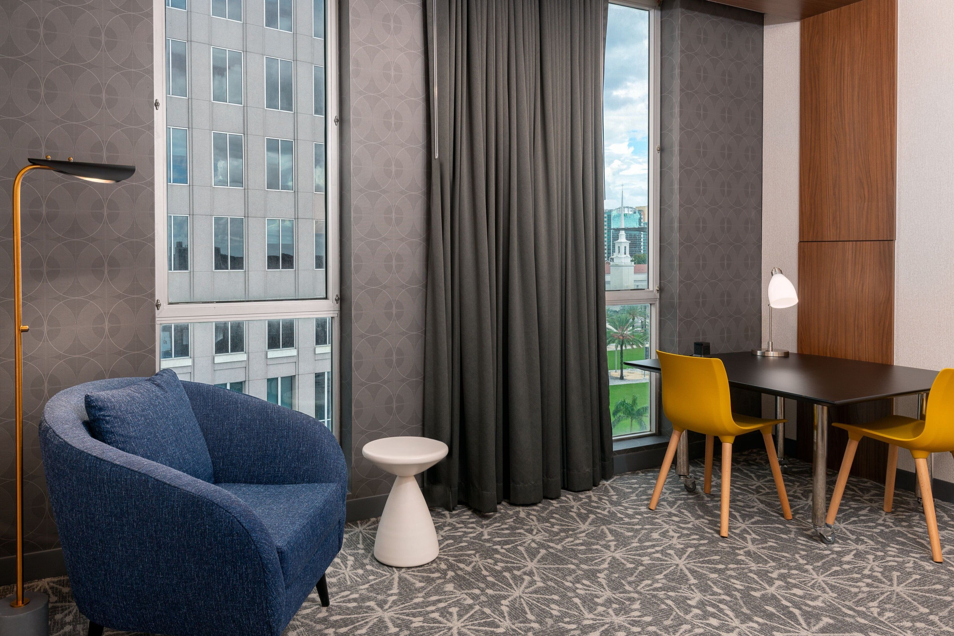Una habitación moderna con una silla azul, una mesa de comedor con sillas amarillas, una mesita auxiliar blanca y vistas a un edificio a través de una ventana con cortinas grises.