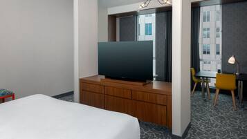 A hotel room with a large bed, a wooden dresser, a flat-screen TV, a desk with a chair, and a window with a view of buildings.