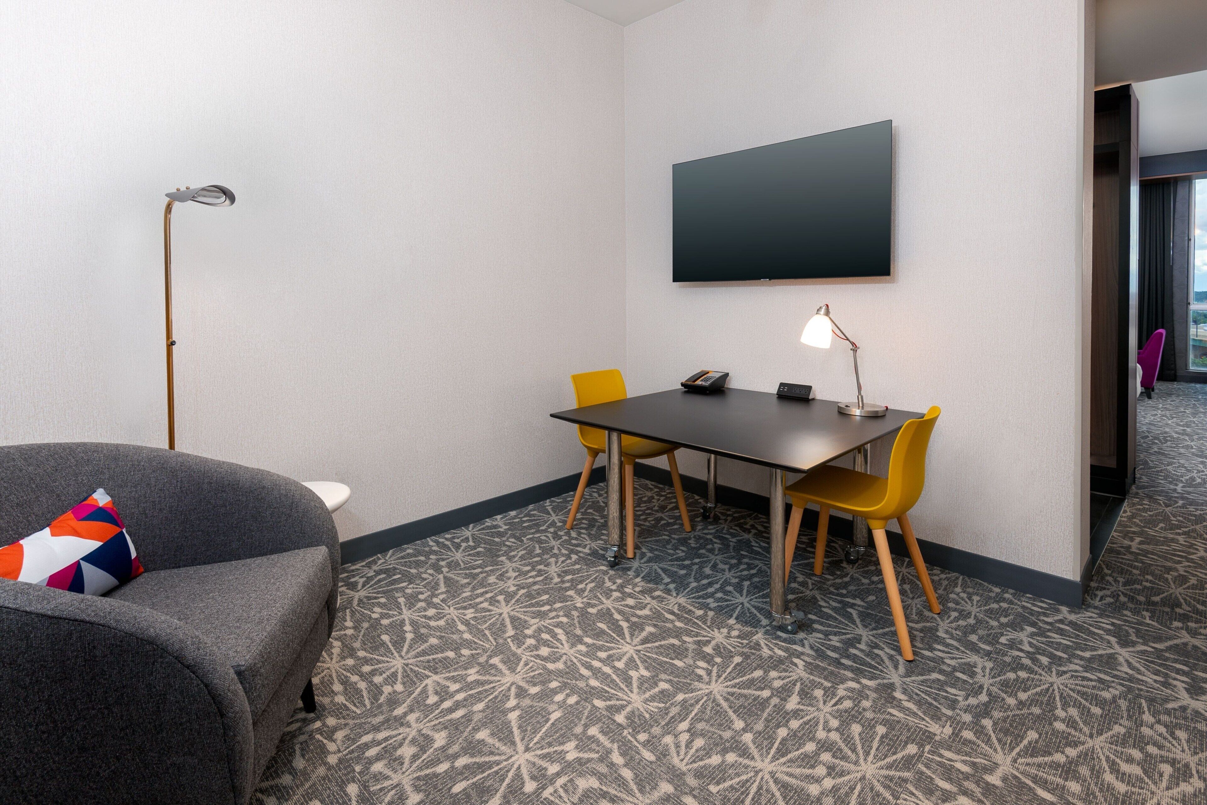 Habitación de hotel con un sofá gris, una mesita pequeña, dos sillas amarillas, un televisor de pantalla plana montado en la pared y alfombra estampada.