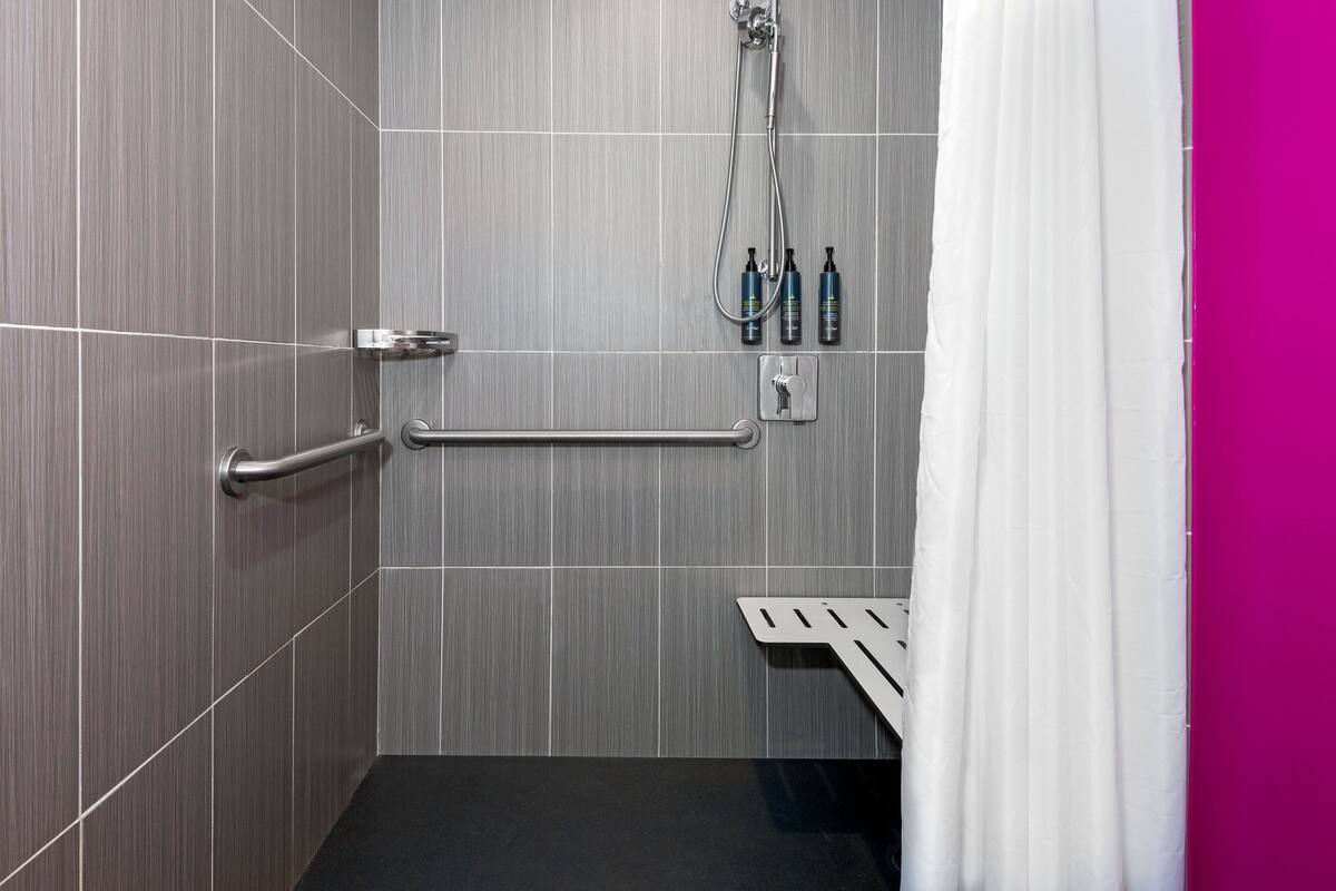 A modern bathroom with grey tiles, a shower with a handheld showerhead, a grab bar, and a shelf with toiletries.