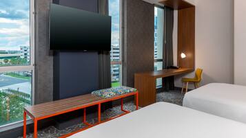A hotel room with a bed, a desk, a chair, a bench, a TV, and a view of the cityscape.