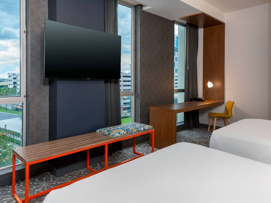 A hotel room with a bed, a desk, a chair, a bench, a TV, and a view of the cityscape.