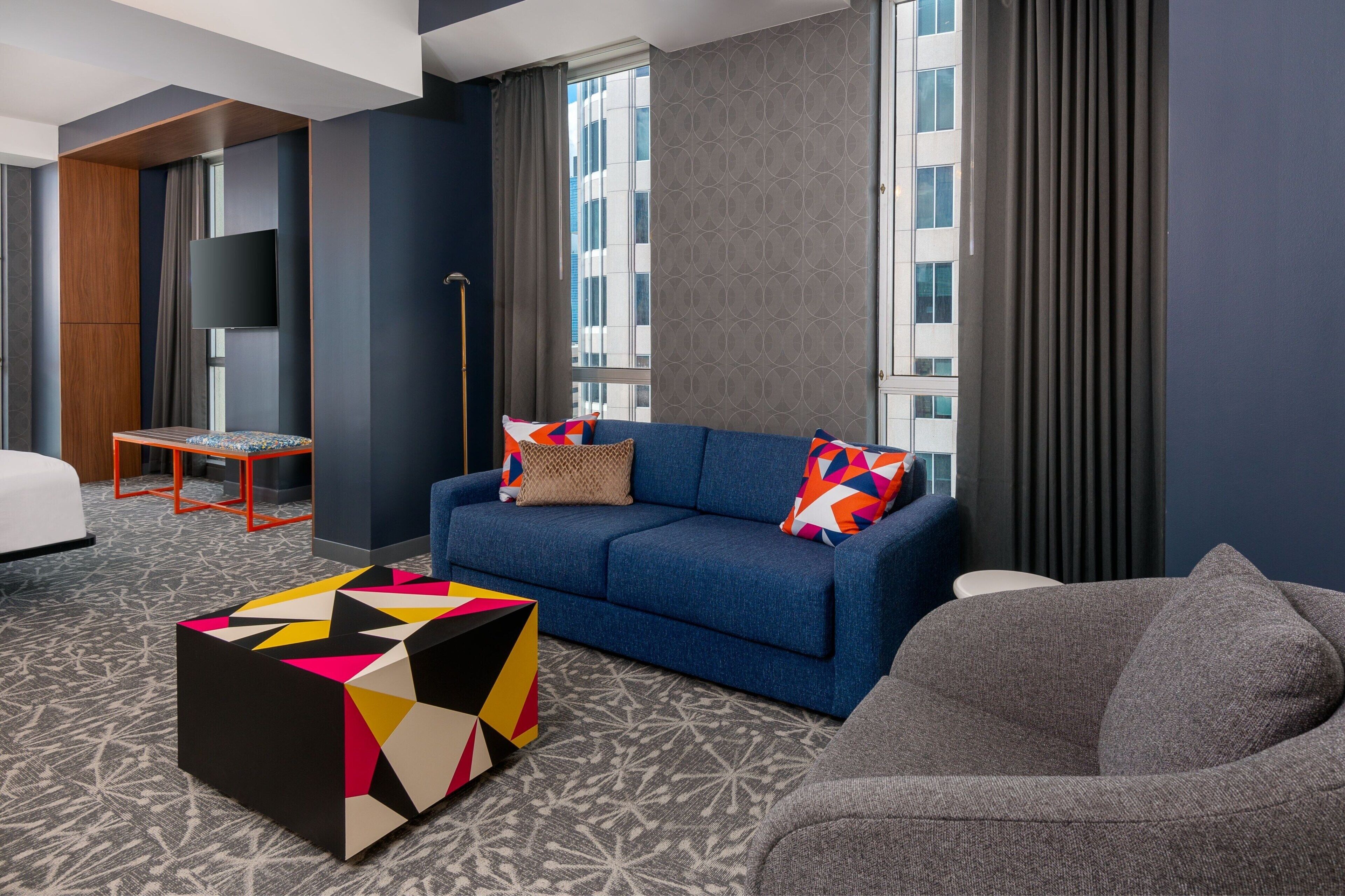 Une chambre d’hôtel moderne avec un canapé bleu, des coussins colorés, une table basse aux motifs géométriques et une moquette à motifs.