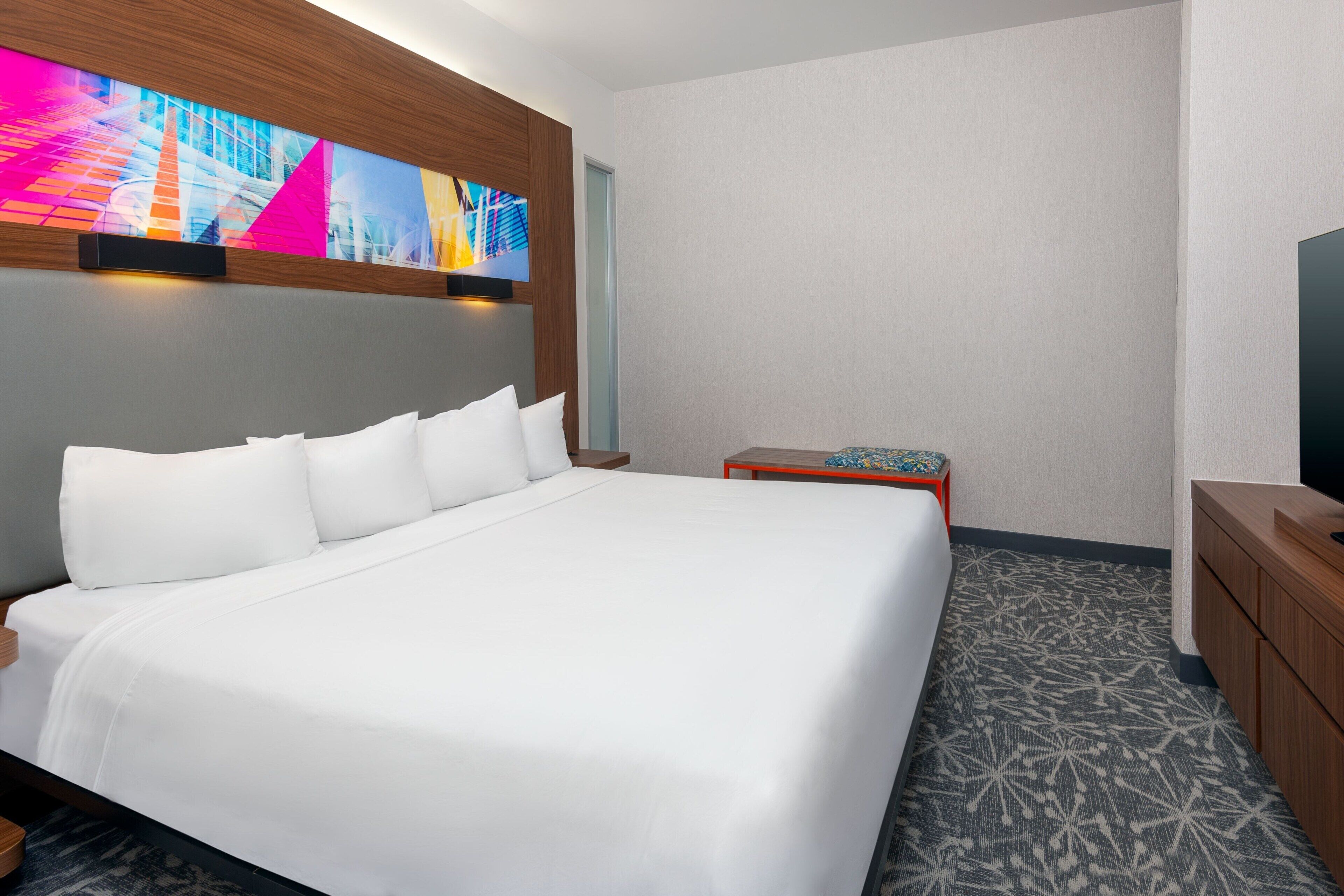A hotel room with a large bed, a small table, a TV, and a wall-mounted artwork.