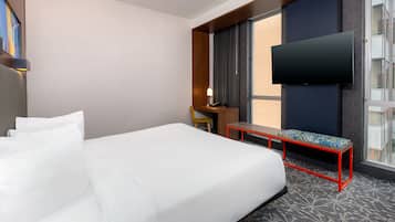 A hotel room with a large bed, a desk with a chair, a bench, a flat-screen TV, and a window with a view.