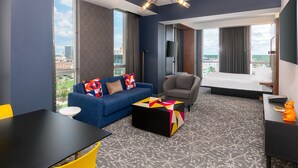 A modern hotel room with a blue sofa, a grey armchair, a black coffee table, and a bed with a white headboard.