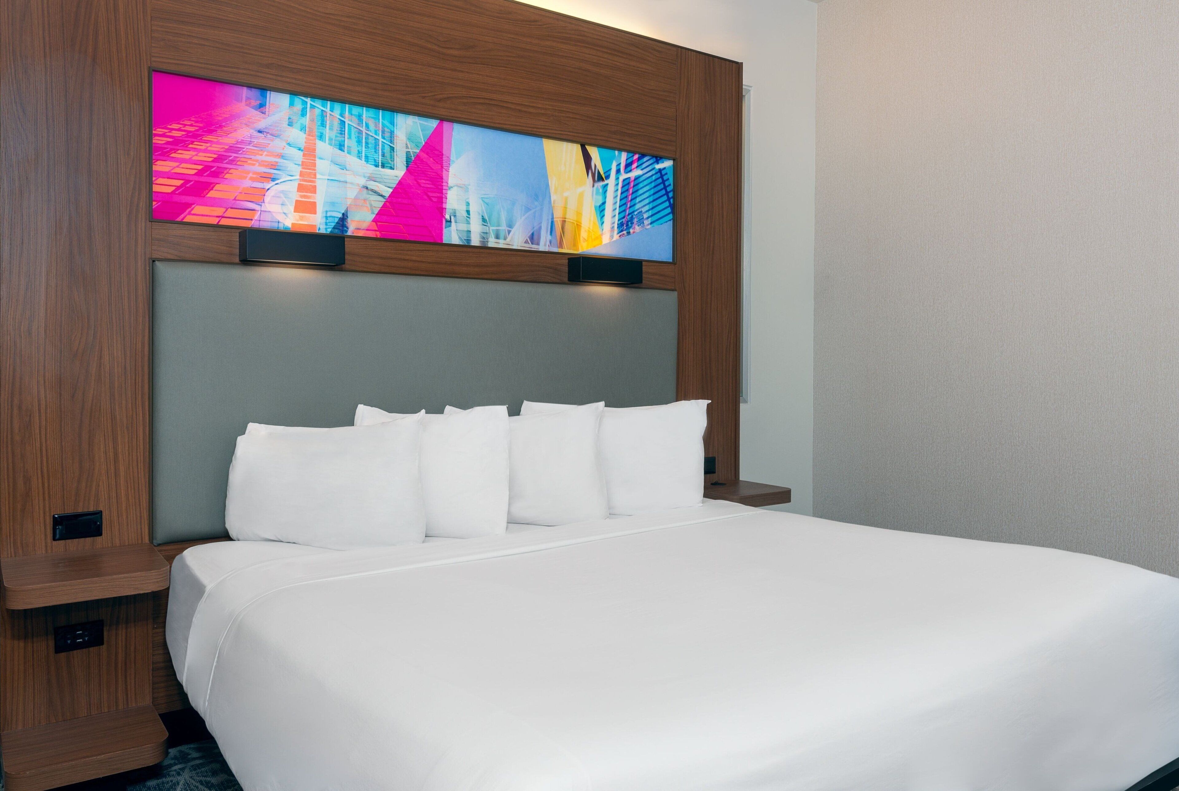 A hotel room with a large bed, white bedding, a wooden headboard, and a colorful artwork above the bed.