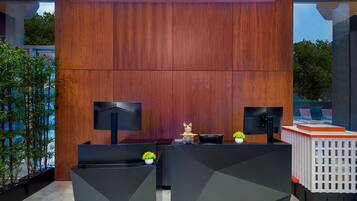 Un mostrador de recepción con fondo de madera, dos monitores, una pequeña estatua decorativa de un gato y una planta en maceta.