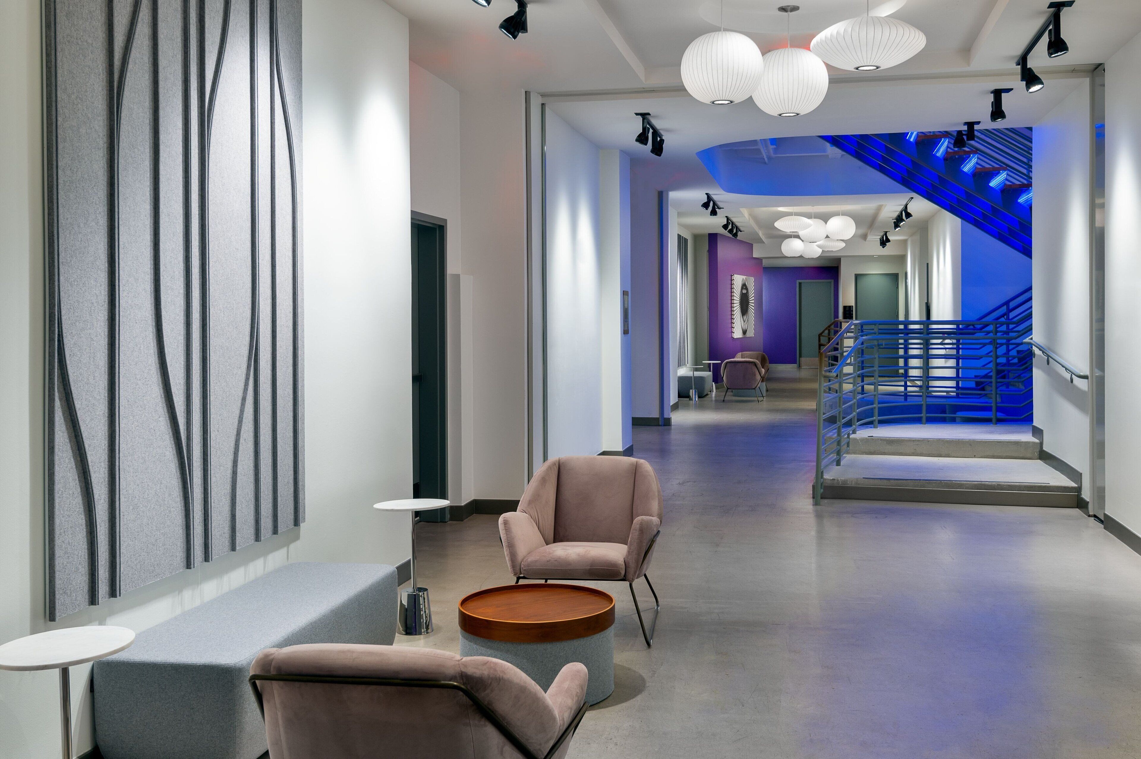 A modern hotel lobby with a central seating area, a staircase with blue lighting, and a view of the hallway leading to other rooms.