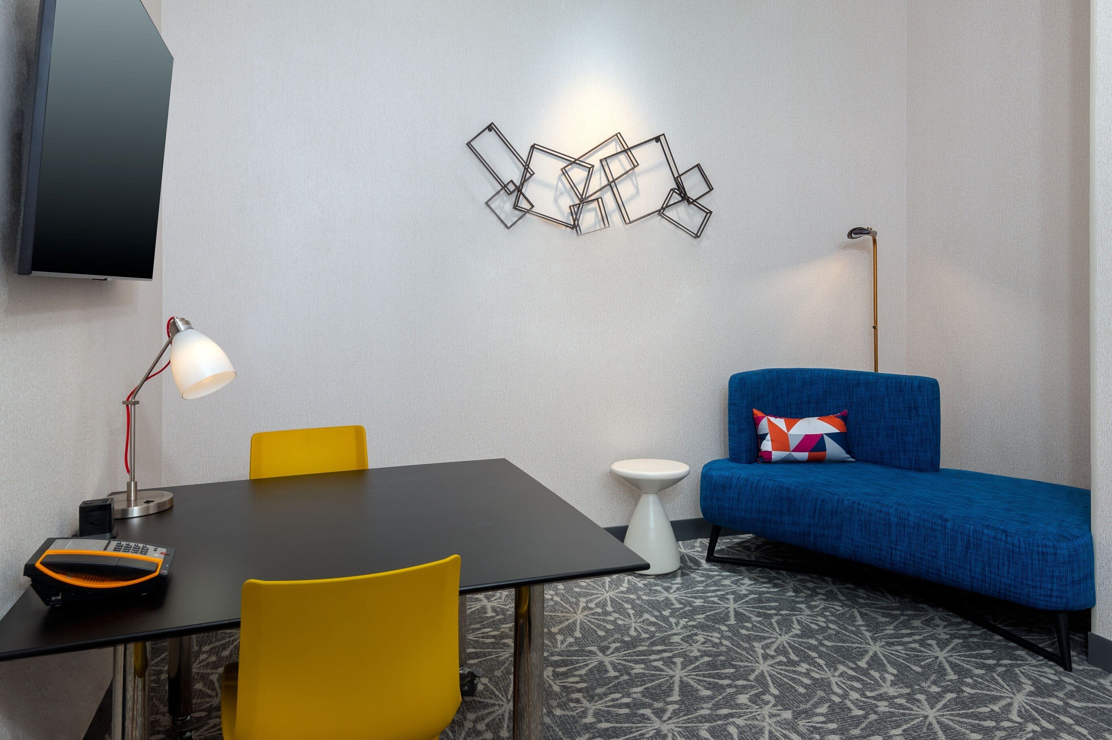 A modern room with a black table, yellow chairs, a blue sofa with a colorful pillow, a white side table, and a wall-mounted TV.