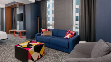 Et moderne hotelværelse med en blå sofa, farverige puder, et geometrisk sofabord og en grå lænestol.