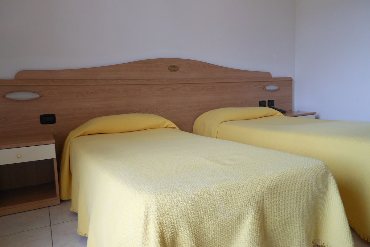 A room with two beds, each with a wooden headboard and yellow bedding. There is a nightstand with a lamp and a small drawer.