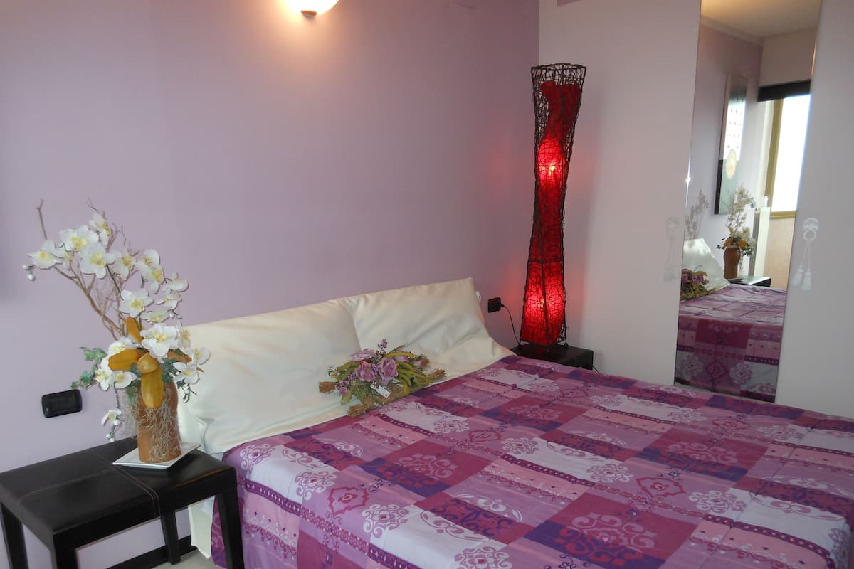 A bedroom with a bed, a nightstand, a vase with flowers, a red lamp, and a mirror.