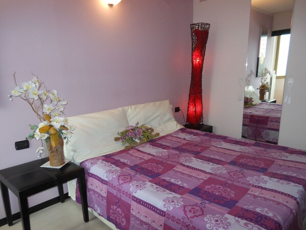 Une chambre à coucher avec un lit, une table de chevet, un vase de fleurs, une lampe rouge et un miroir.