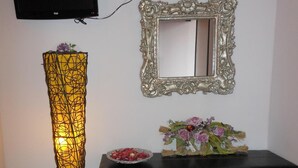A room with a TV mounted on the wall, a decorative mirror, a black console table with drawers, a small stool, and a tall, yellow, wireframe lamp. - Motel Monterosa (Vogogna)