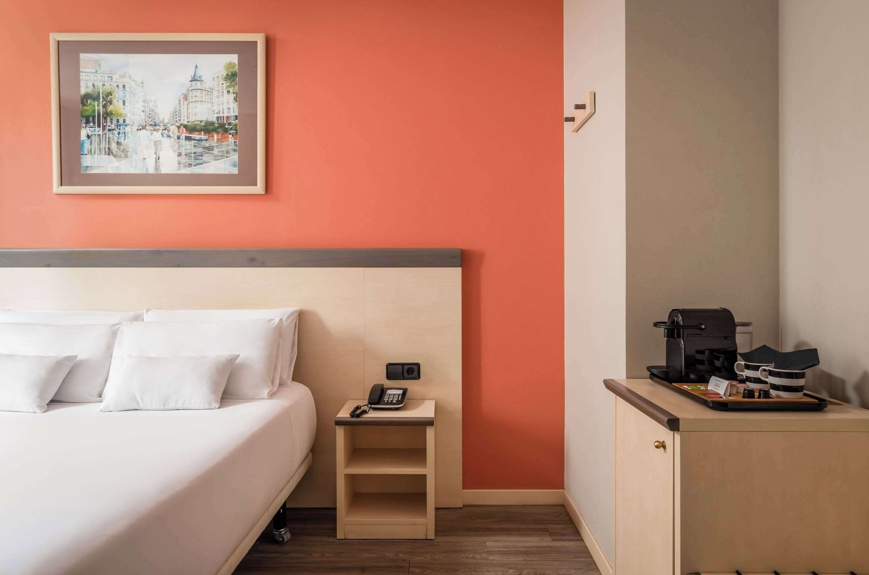 A hotel room with a bed, a nightstand with a phone, a coffee machine, and a framed cityscape painting on the wall.