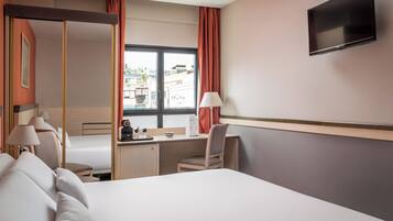 Una habitación de hotel con cama, escritorio, silla, televisor y ventana con cortinas.