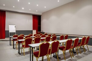 Un salón de conferencias con filas de mesas y sillas, una pizarra blanca y un telón rojo de fondo.