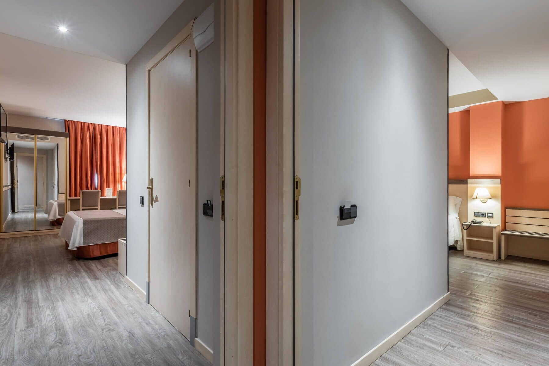 A hotel room with a bed, a desk, and a chair. There is a door with a handle and a light switch on the wall.