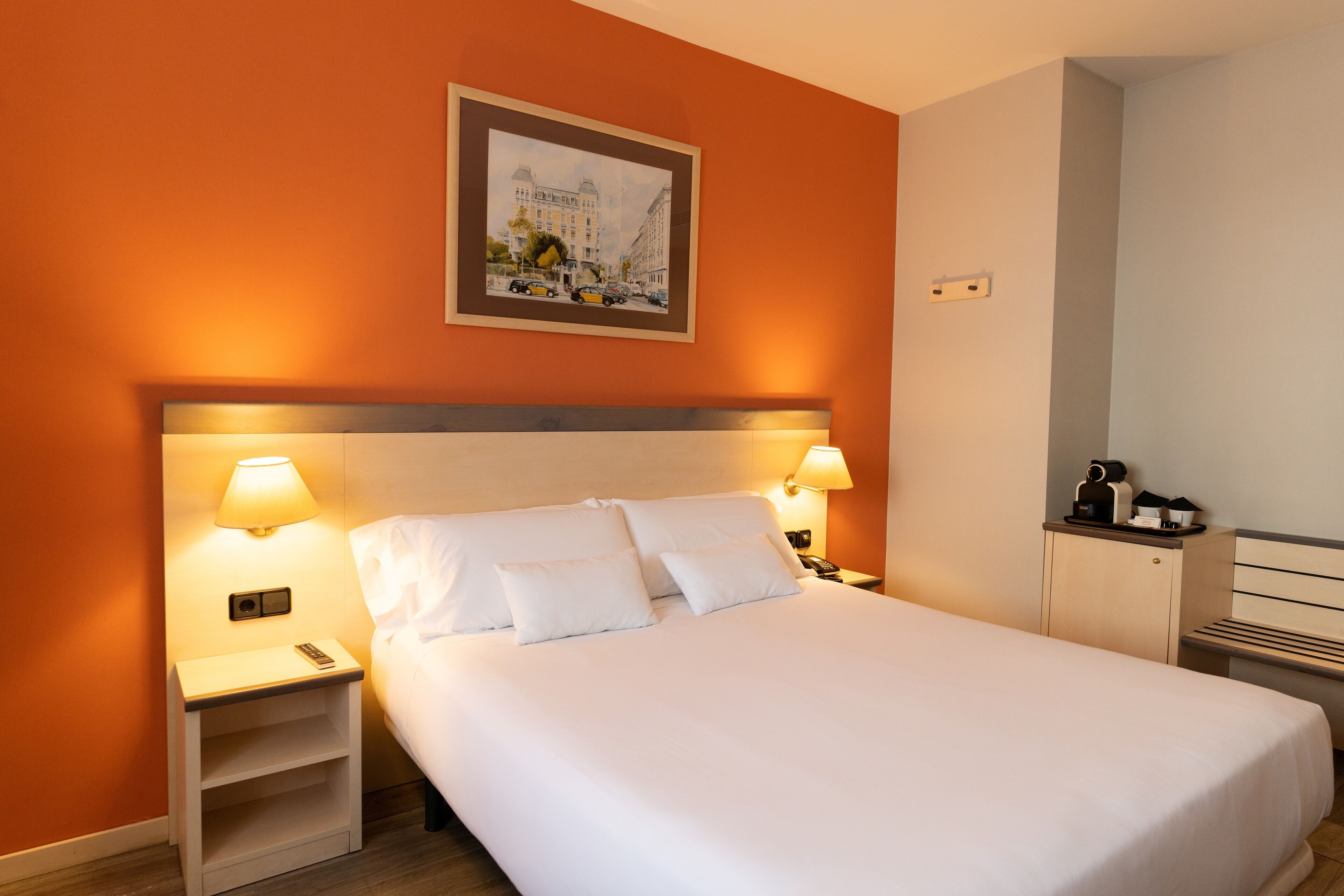 Hotellihuone, jossa on suuri sänky, kaksi yöpöytää valaisimineen, seinällä oleva kehystetty taulu ja pieni kaappi.