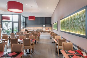 Un restaurante moderno con mesas y sillas altas, una cocina al fondo y una gran obra de arte en la pared.