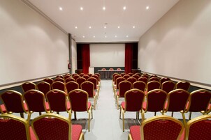 Un salón de conferencias con filas de sillas rojas y un escenario con una pantalla de proyector.