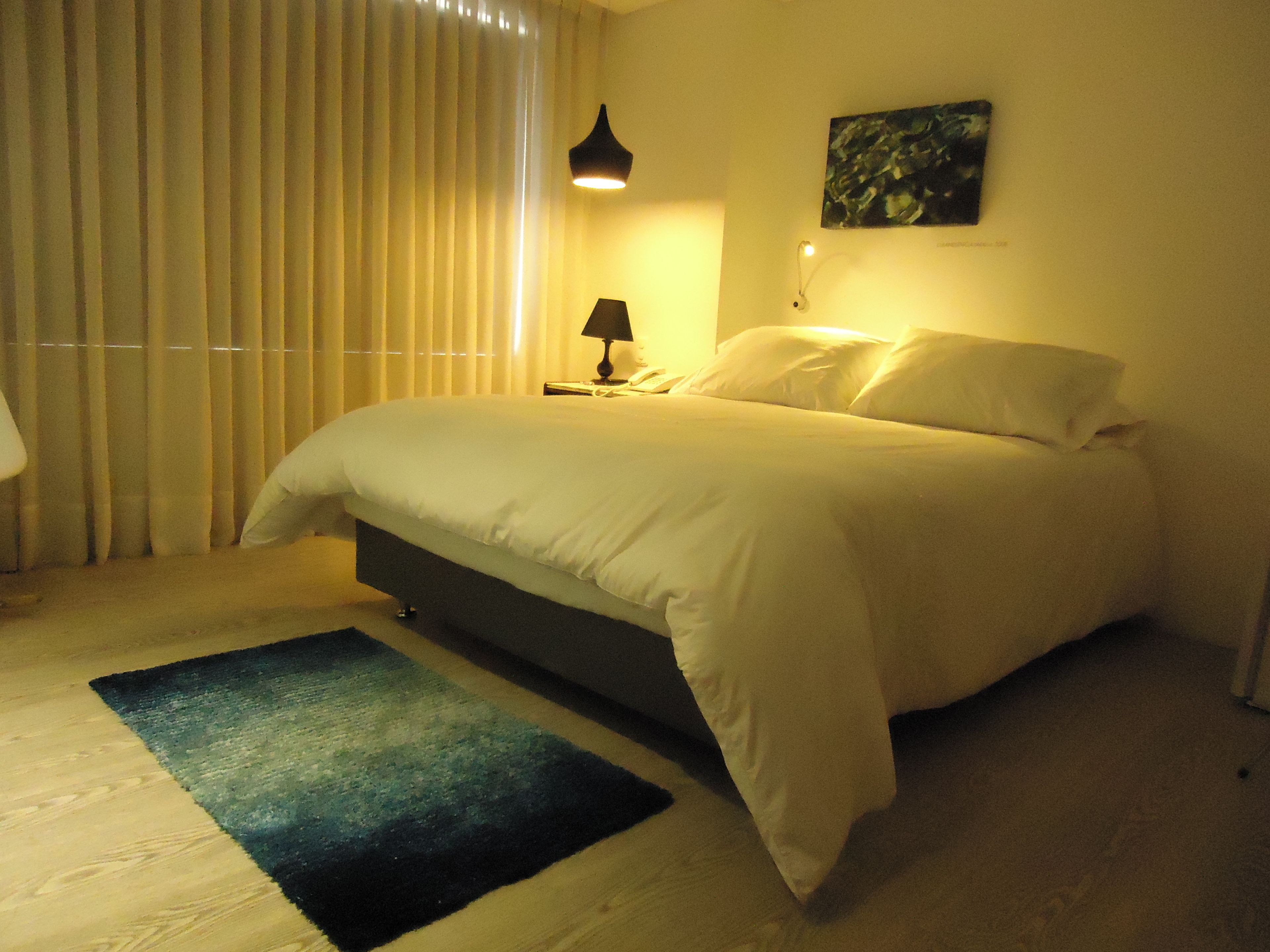 A neatly made bed in a room with a dark-colored rug, a bedside lamp, and a painting on the wall.