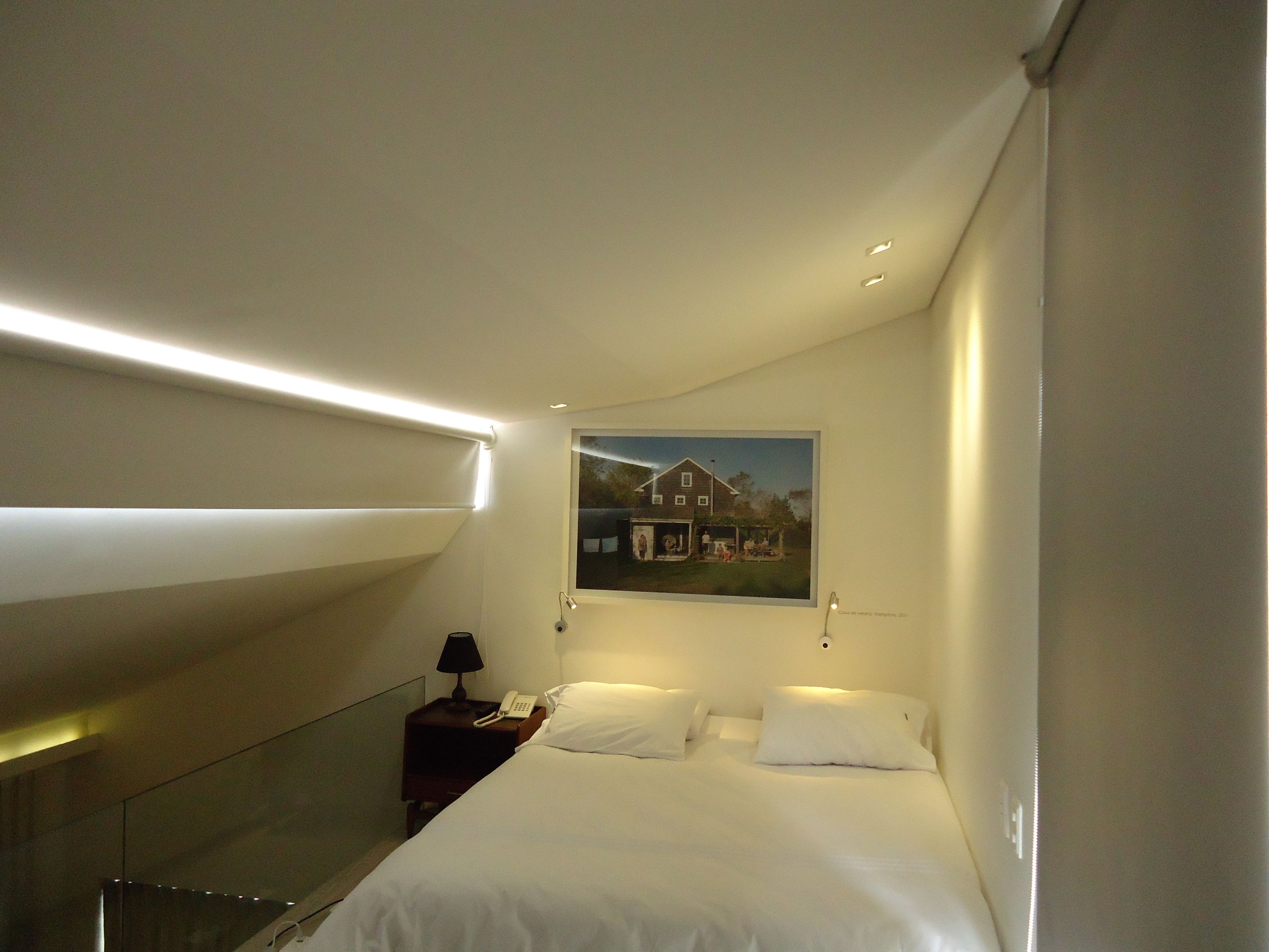 A hotel room with a bed, a nightstand with a lamp and phone, a painting on the wall, and a glass partition.